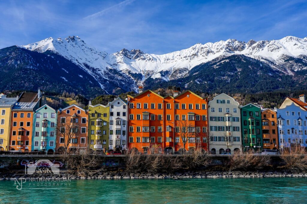 Painted buildings of Innsbruck Austria next to river in front of snowy mountains nathan jordan photography